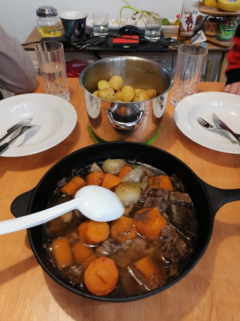 Kuvassa taustalla lautaset ja perunakattila sekä etualalla valurautapata, jossa porkkanoita, lihaa ja sipulia. Padassa on valkoinen kauha.