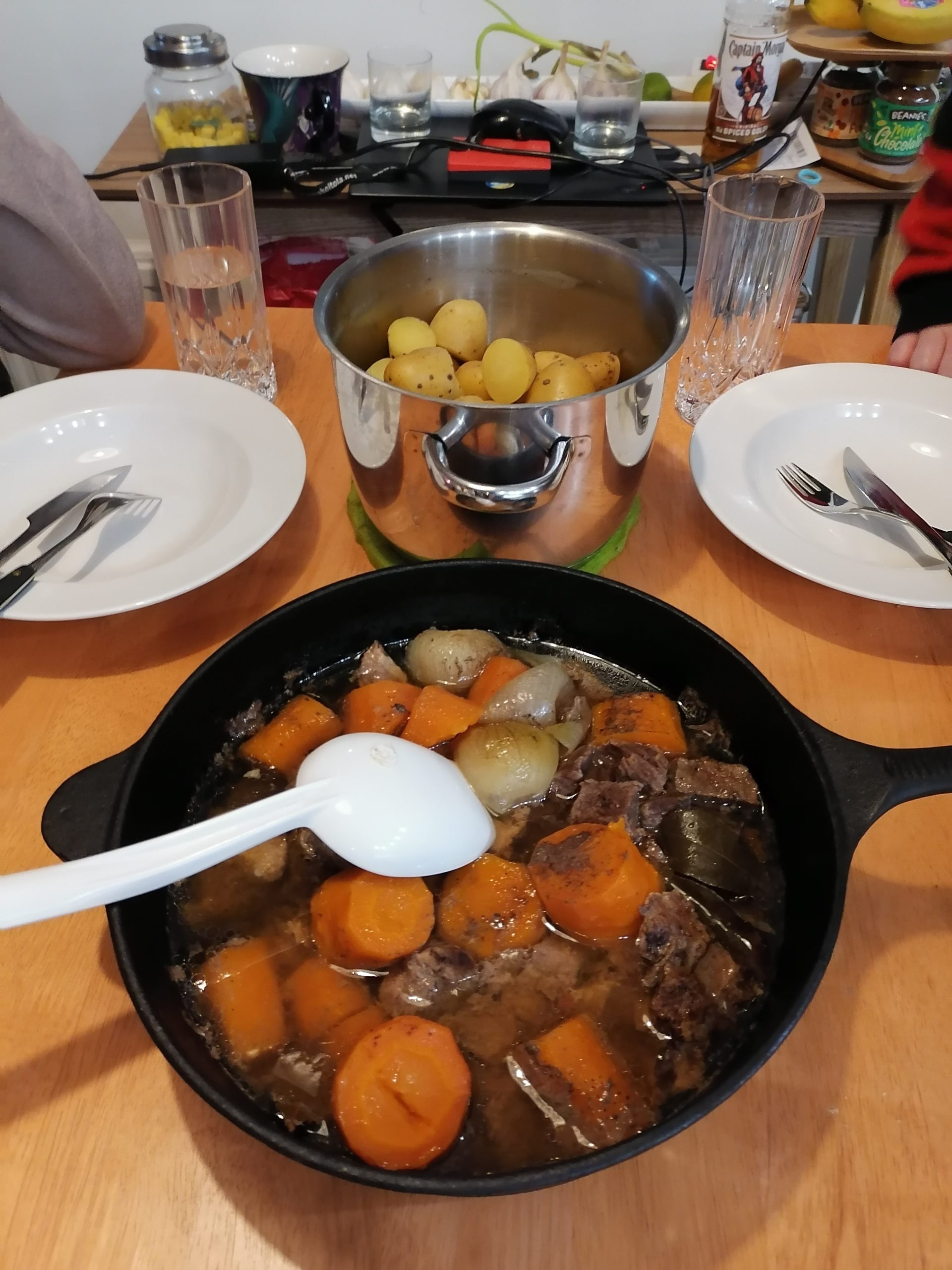 Kuvassa taustalla lautaset ja perunakattila sekä etualalla valurautapata, jossa porkkanoita, lihaa ja sipulia. Padassa on valkoinen kauha.